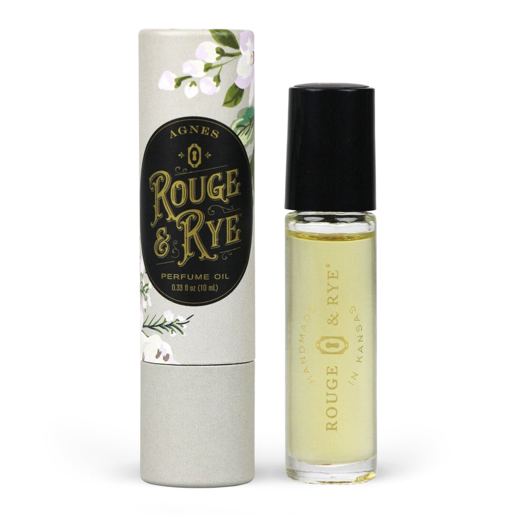 Golden Agnes perfume oil roller in clear glass bottle with black plastic cap next to gray floral packaging. White background.