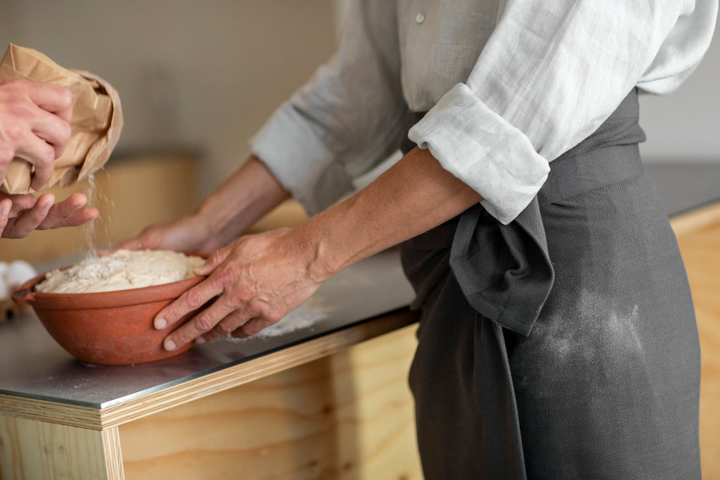 Lifestyle photo of man wearing dark grey towel as an apron while working with a bowl of flour on top of a wood and stainless steel work table. 