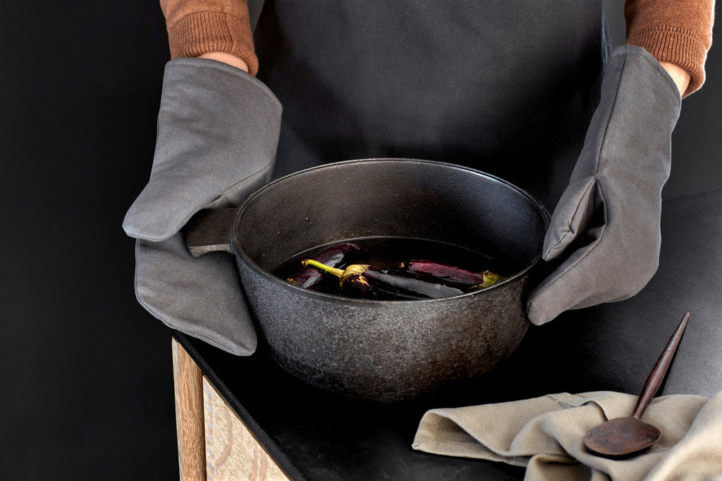 Lifestyle photo of dark gray organic cotton oven mitts on a person wearing a brown sweater and black apron holding a black pot of eggplants. Black countertop with tan napkin and wood spoon.