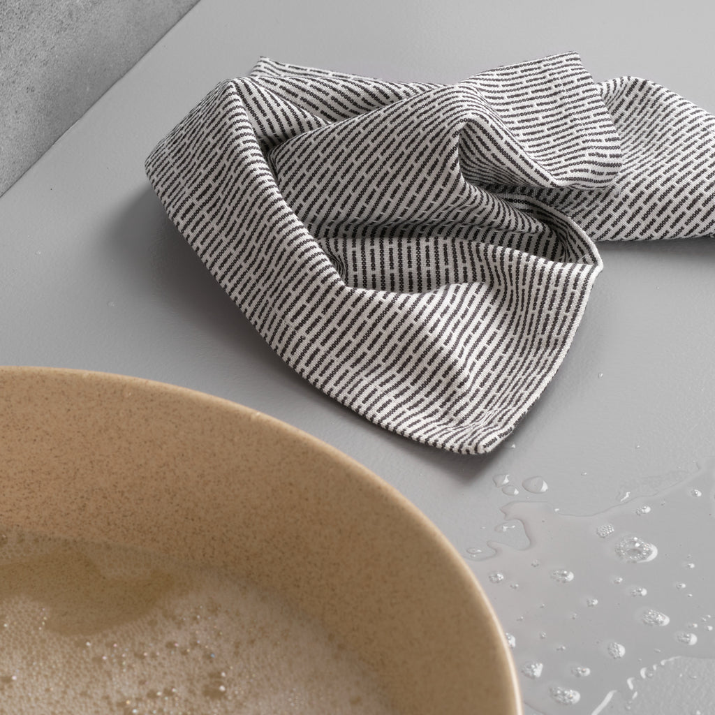 Organic cotton pique woven kitchen and washcloth draped next to a tan bowl of water on a gray countertop. Light gray and cream subtle striping.