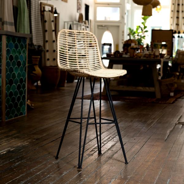 Rattan stripe pattern bar stool sets atop black metal connected hairpin legs. Sits in front of a shop background.