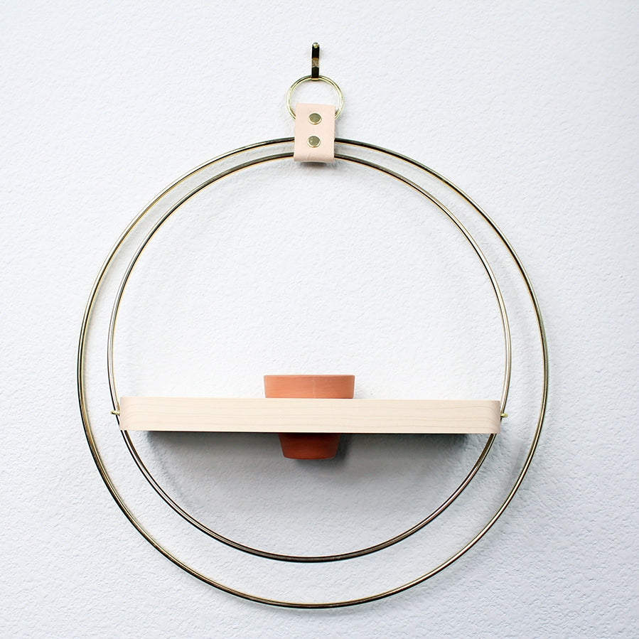 Two large brass rings hold an oak shelf in the middle. The oak shelf has a hole for a small terra cotta pot. Hangs by a leather strap and brass ring at top. White wall behind.