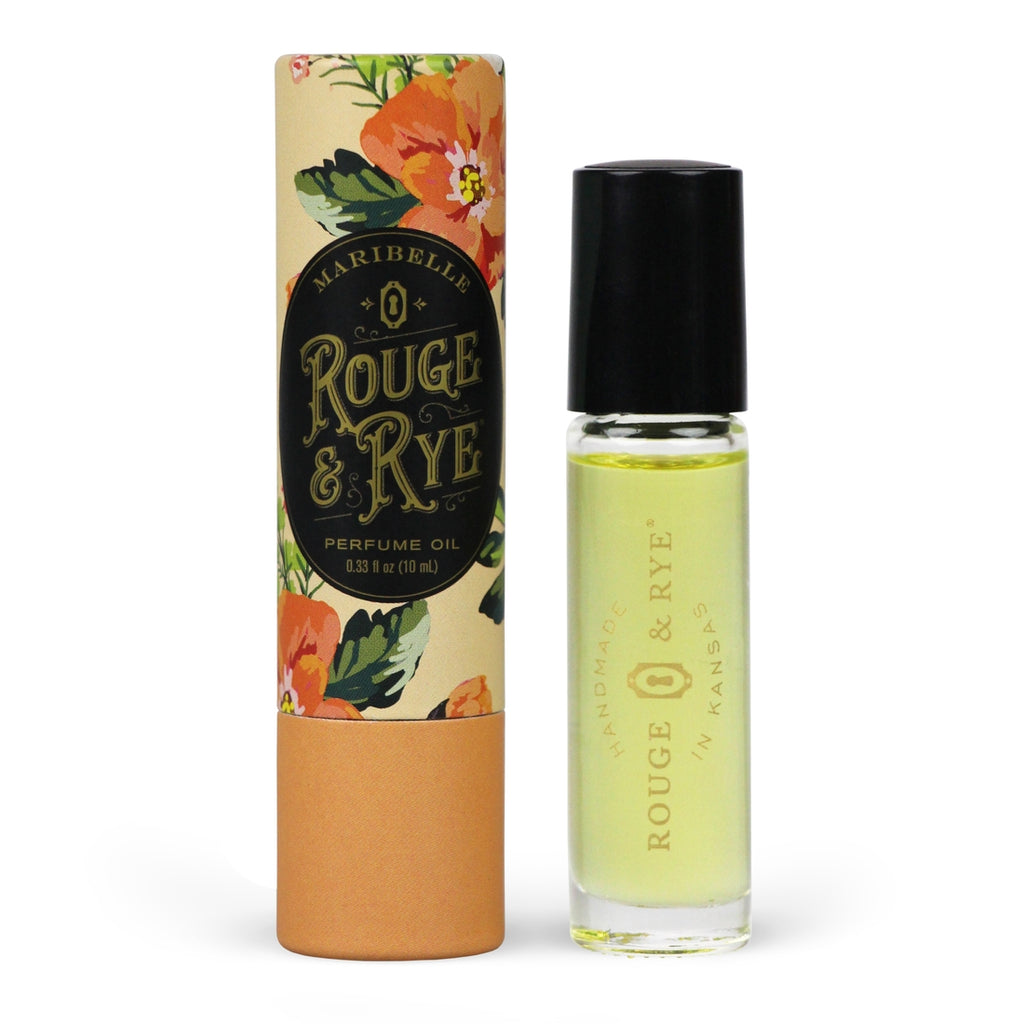 Golden Maribelle perfume oil roller in clear glass bottle with black plastic cap next to orange tropical floral packaging. White background.
