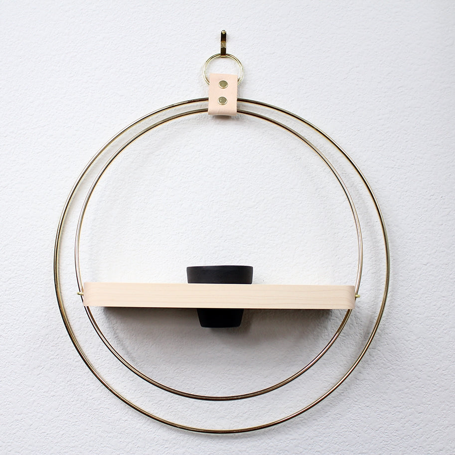 Two large brass rings hold an oak shelf in the middle. The oak shelf has a hole for a small black pot. Hangs by a leather strap and brass ring at top. White wall behind.