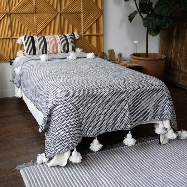 Large cotton blanket woven in a black and white small diamond pattern with big white pom poms along the edge. Laying on a twin sized bed.