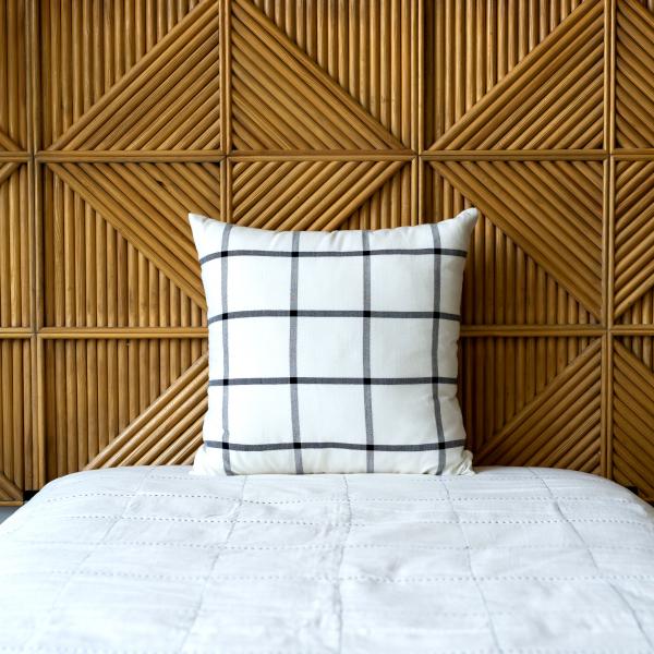 Cream woven cotton pillow features a black grid pattern. Sits on a bed with a white quilt and a wood headboard.