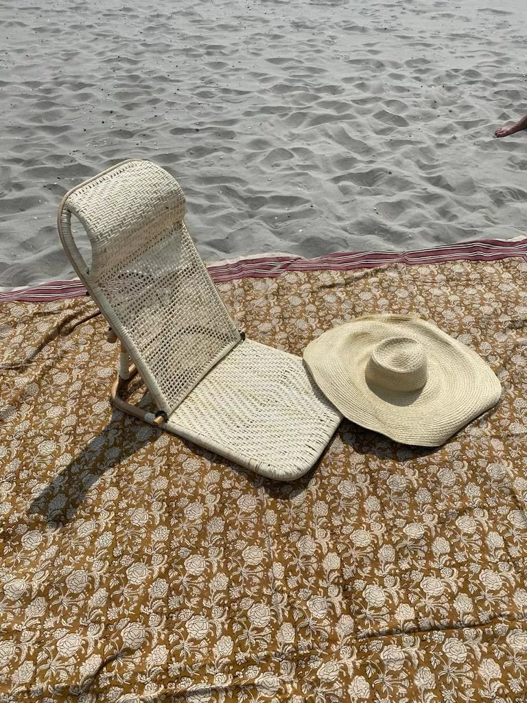 Foldable rattan park chair sits on a blanket next to sand. A sun hat sits next to the chair sitting on the ground.