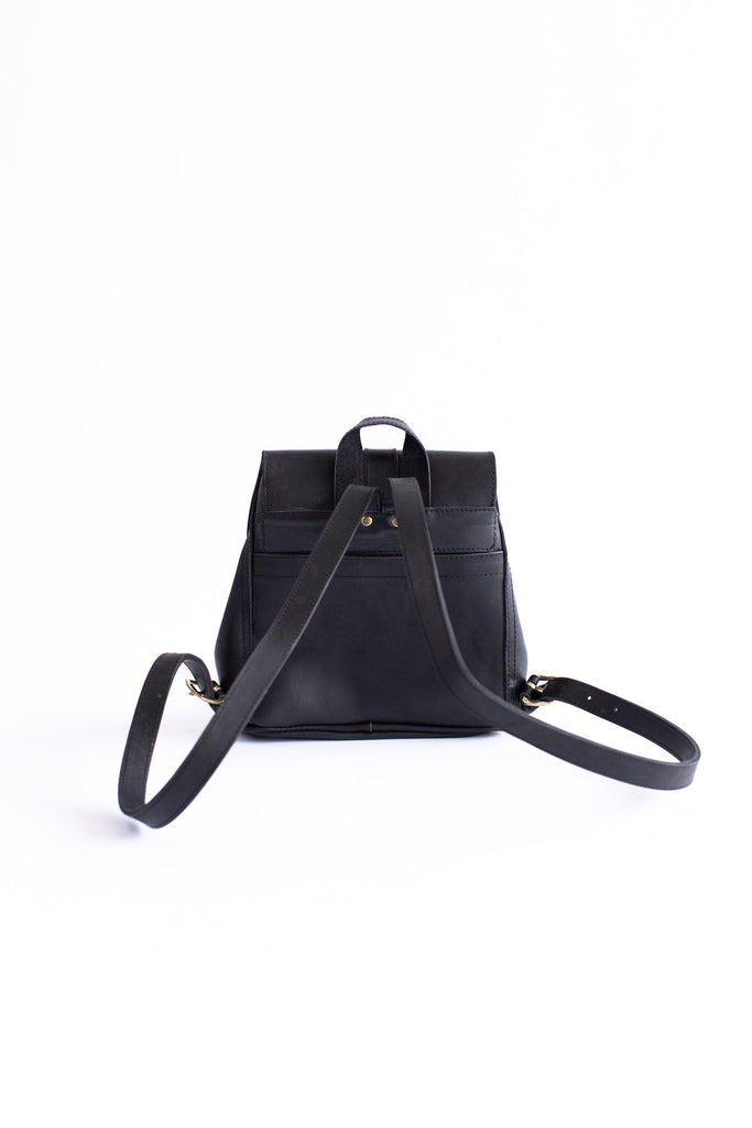 The back of a vegetable tanned black leather small backpack sits on a white background. Hanging loop and adjustable straps are shown.