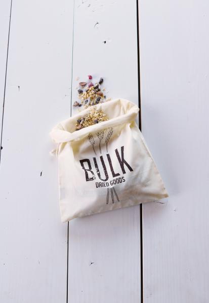 Organic cotton drawstring produce bag. Design says “Bulk Dried Goods” in brown ink. The text is in front of 3 illustrated wheat grains. Pictured with the top rolled down and grains spilling out.