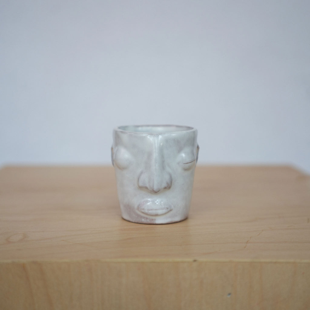 One white ceramic face mezcal cups sit on a wood platform in front of a white background. 