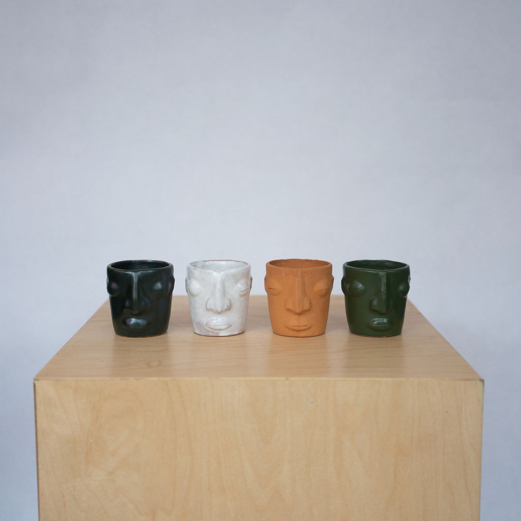 Group of four ceramic face mezcal cups sit on a wood platform in front of a white background. Green gloss, white, terra cotta, and matte green.