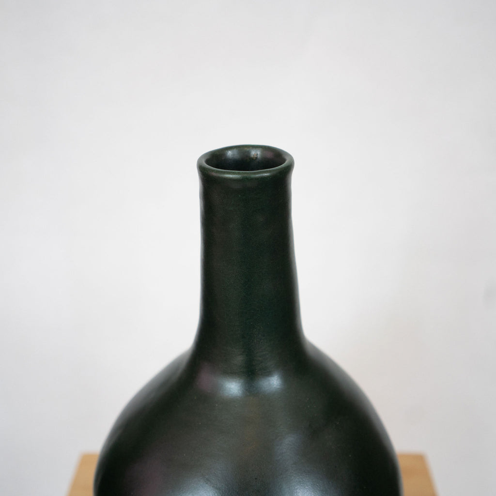 Dark green ceramic vase with a round bottom and slim column neck. Sits on a wood platform with a white background.