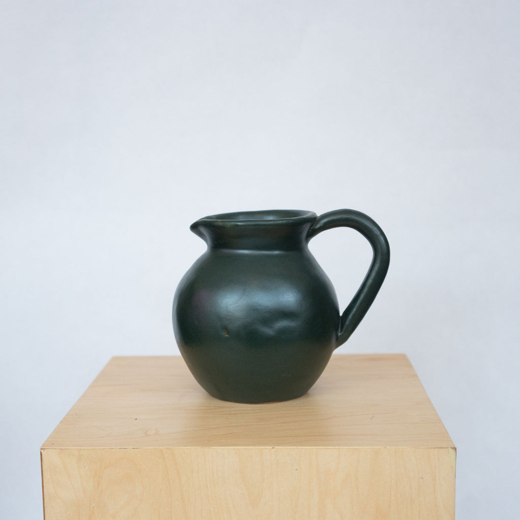 Dark green ceramic pitcher on wood pedestal in front of a white background.