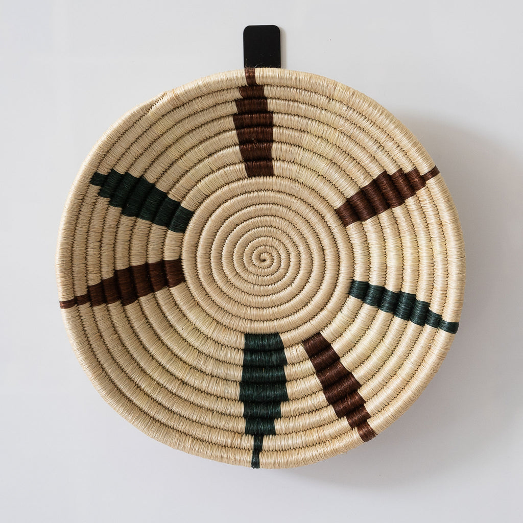 Handwoven Rwandan sweetgrass basket bowl with a slightly abstract petal design in tan, brown, and dark green. White background.