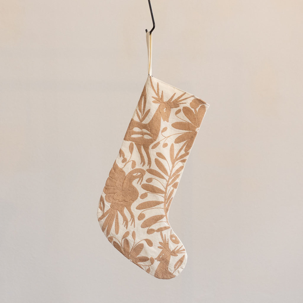 Holiday / Christmas stocking in traditional Otomi embroidered style with tan flora and fauna on a cream background. Hangs from a black S-hook in front of a tan gray background.