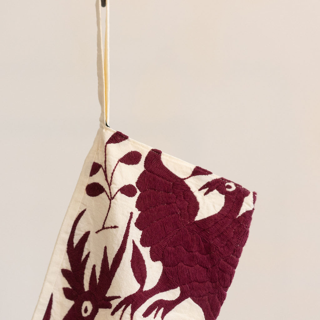 Holiday / Christmas stocking in traditional Otomi embroidered style with maroon flora and fauna on a cream background. Hangs from a black S-hook in front of a tan gray background.