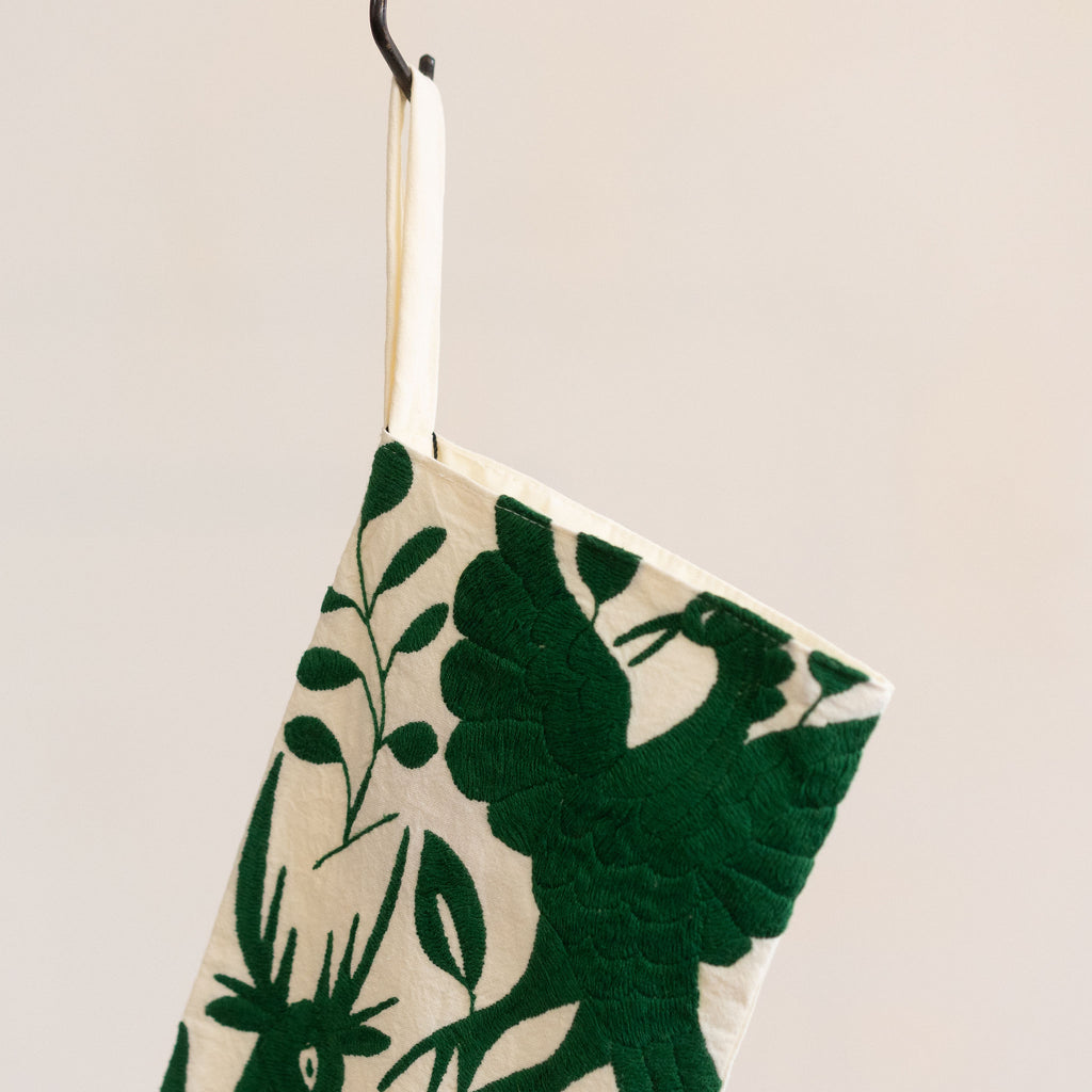 Holiday / Christmas stocking in traditional Otomi embroidered style with green flora and fauna on a cream background. Hangs from a black S-hook in front of a tan gray background.