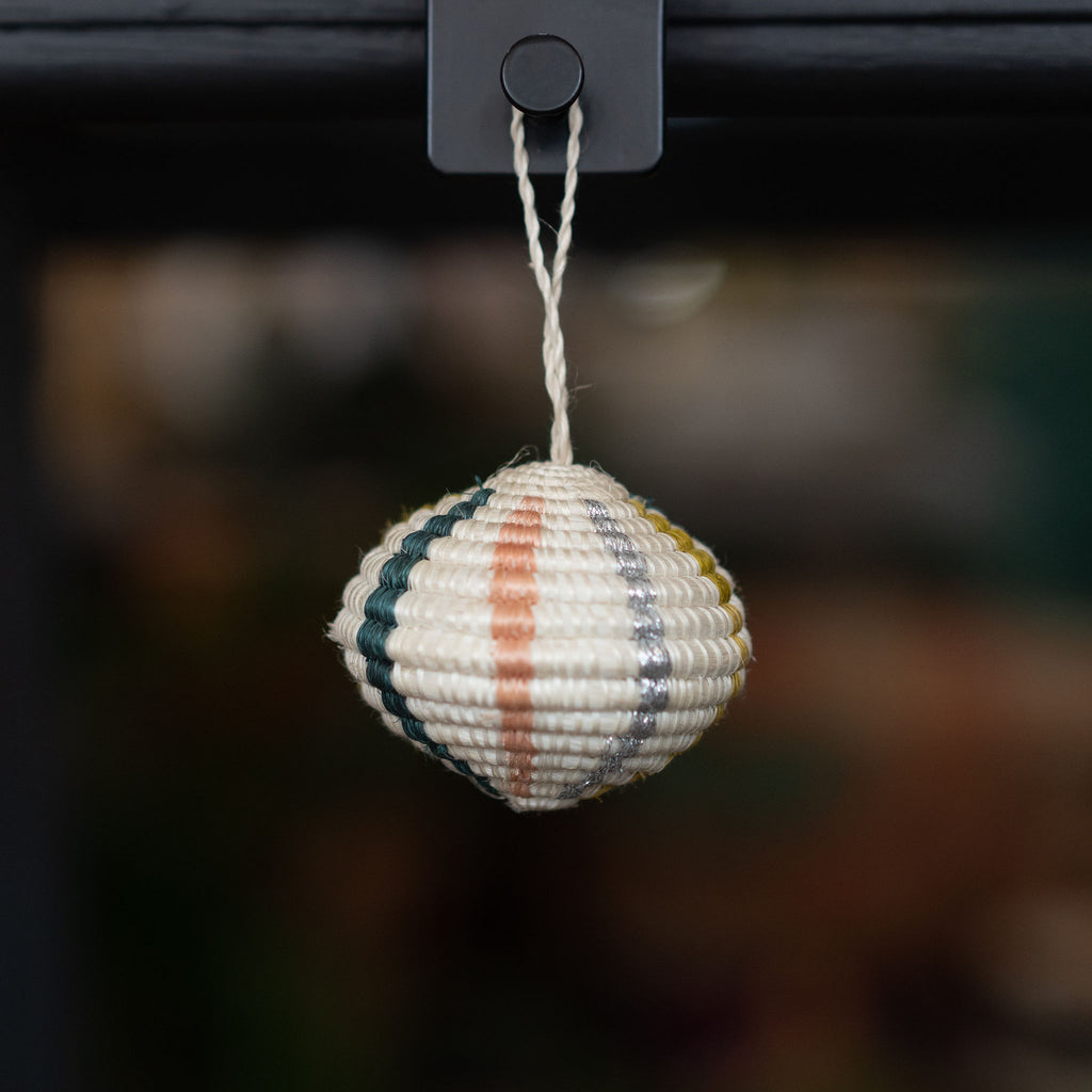 Handwoven round striped sweetgrass Rwandan tree ornament in cream, blush, silver, mustard, and dark green. Blurry shop background.