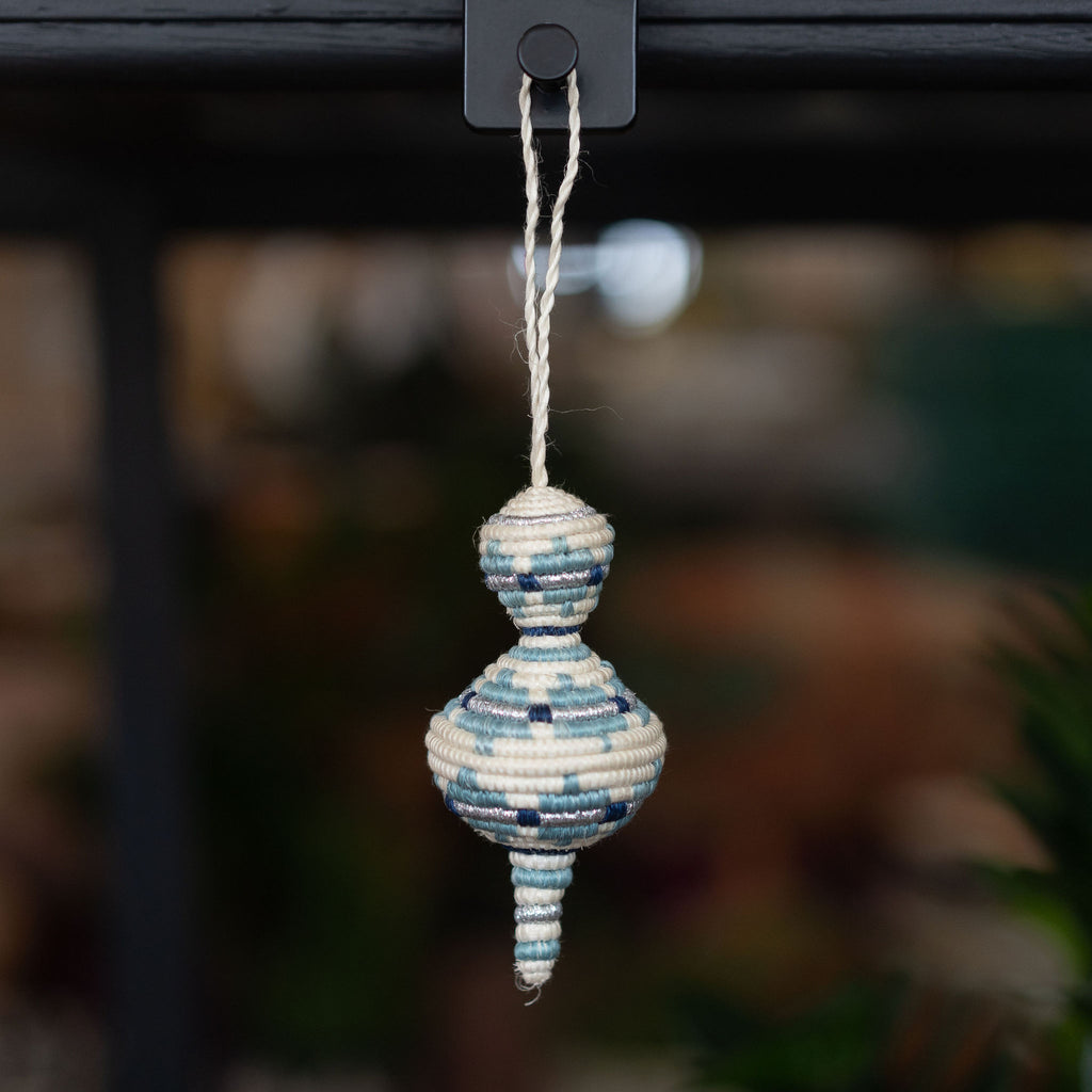 Handwoven sweetgrass Rwandan tree ornament shaped like a curvy lantern in shades of blue with silver and cream. Blurry shop background. 