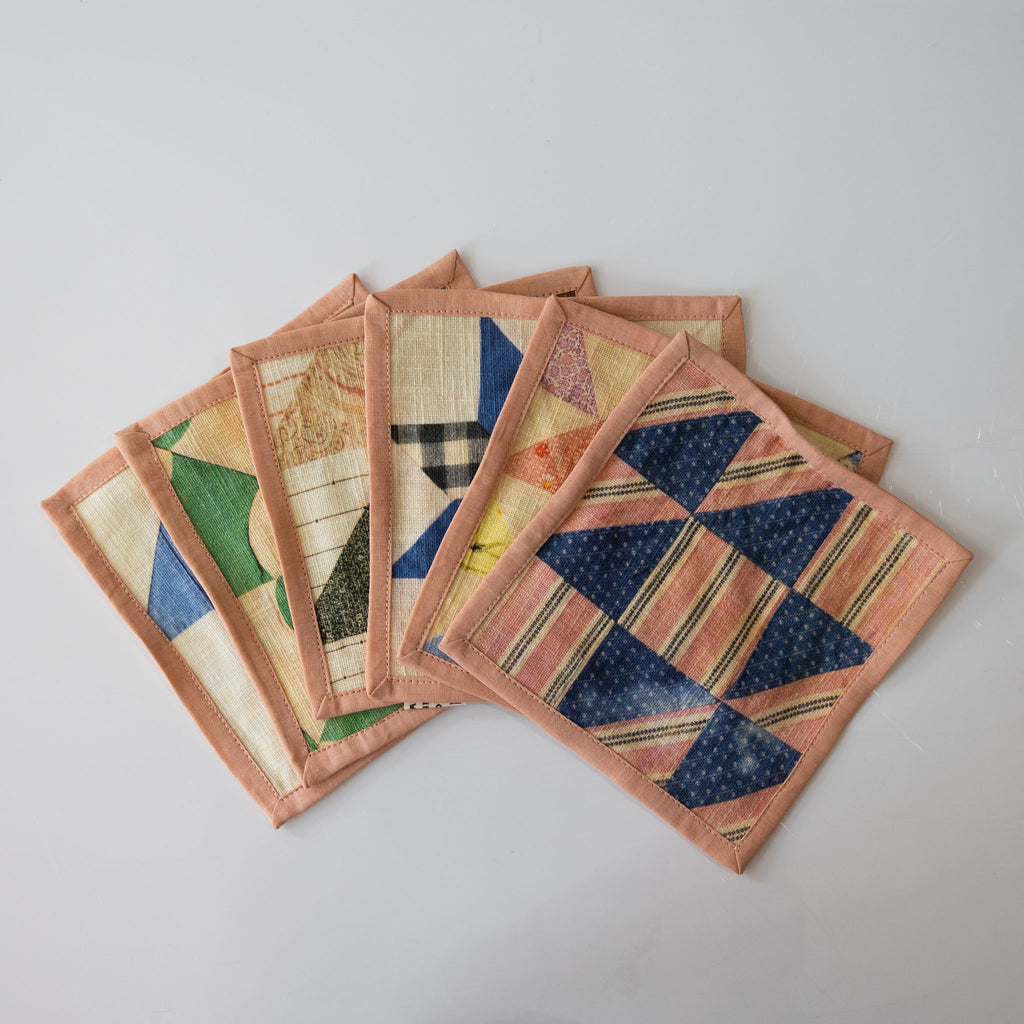 A fanned stack of six assorted printed quilt patterned cocktail napkins in coral, yellow, blue, green, and cream. White background.