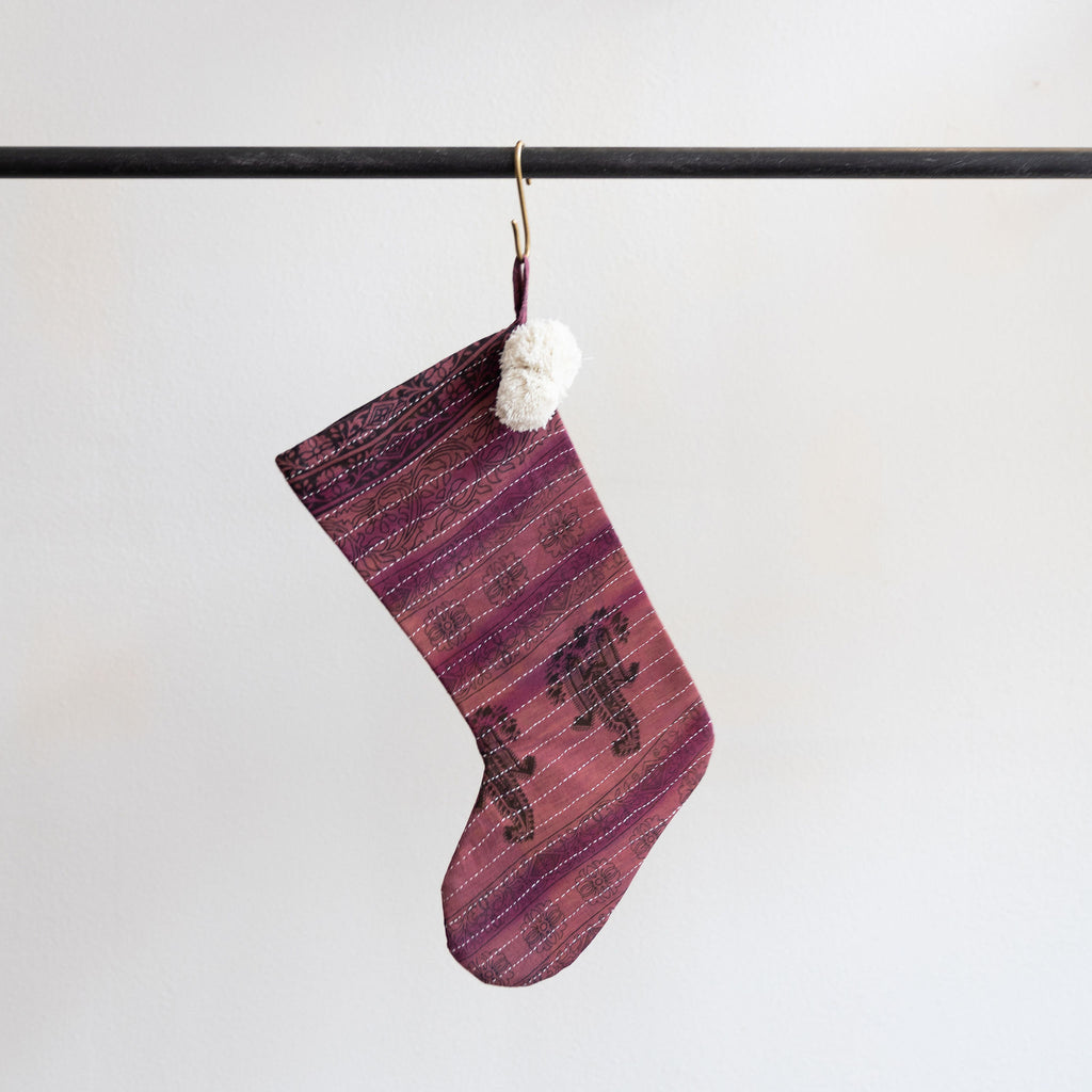 A holiday stocking that is handmade and embroidered with the traditional Kantha stitch. Shades of pink and purple recycled sari fabric and cream poms by the hanging loop. Hanging from a brass s-hook from a black bar in front a white background.  
