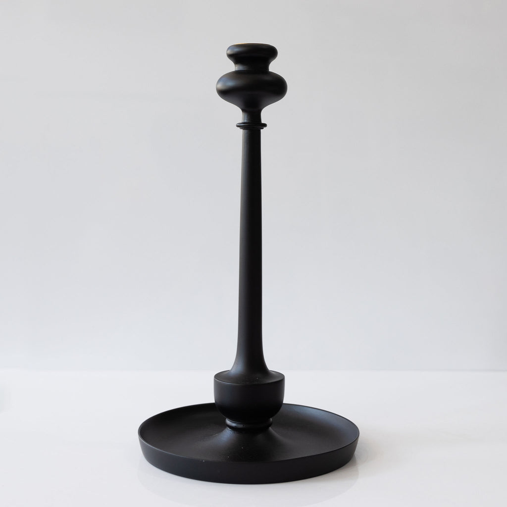 A tall curvy wood taper candlestick with a wide tray at the bottom. Matte black. Sits against a white background.