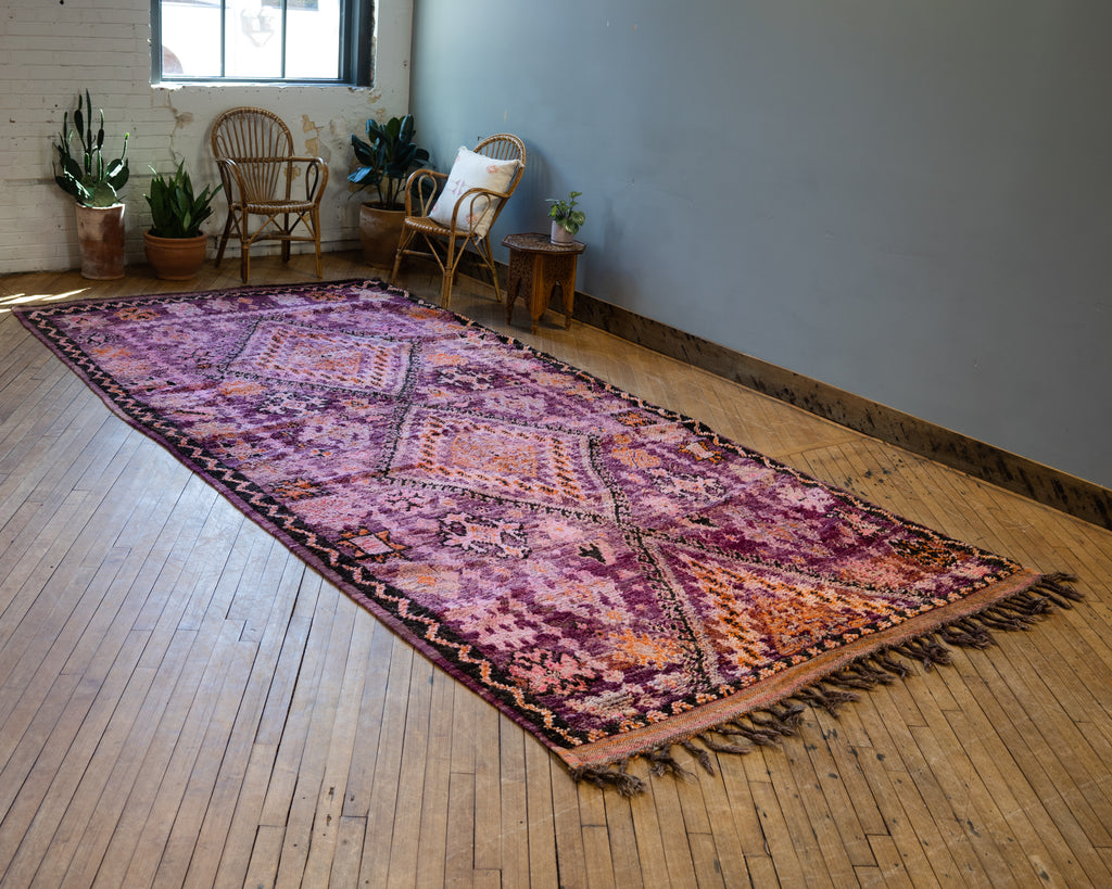 A diagonal view of a vintage Moroccan Boujad rug with a bold diamond design in shades of purple, orange, and pink. Two diamonds are surrounded by various Berber symbols. A zig zag border surrounds an edge section with repeat Berber symbols. Surrounded by two rattan chairs, a side table, and potted plants. Wood floors.