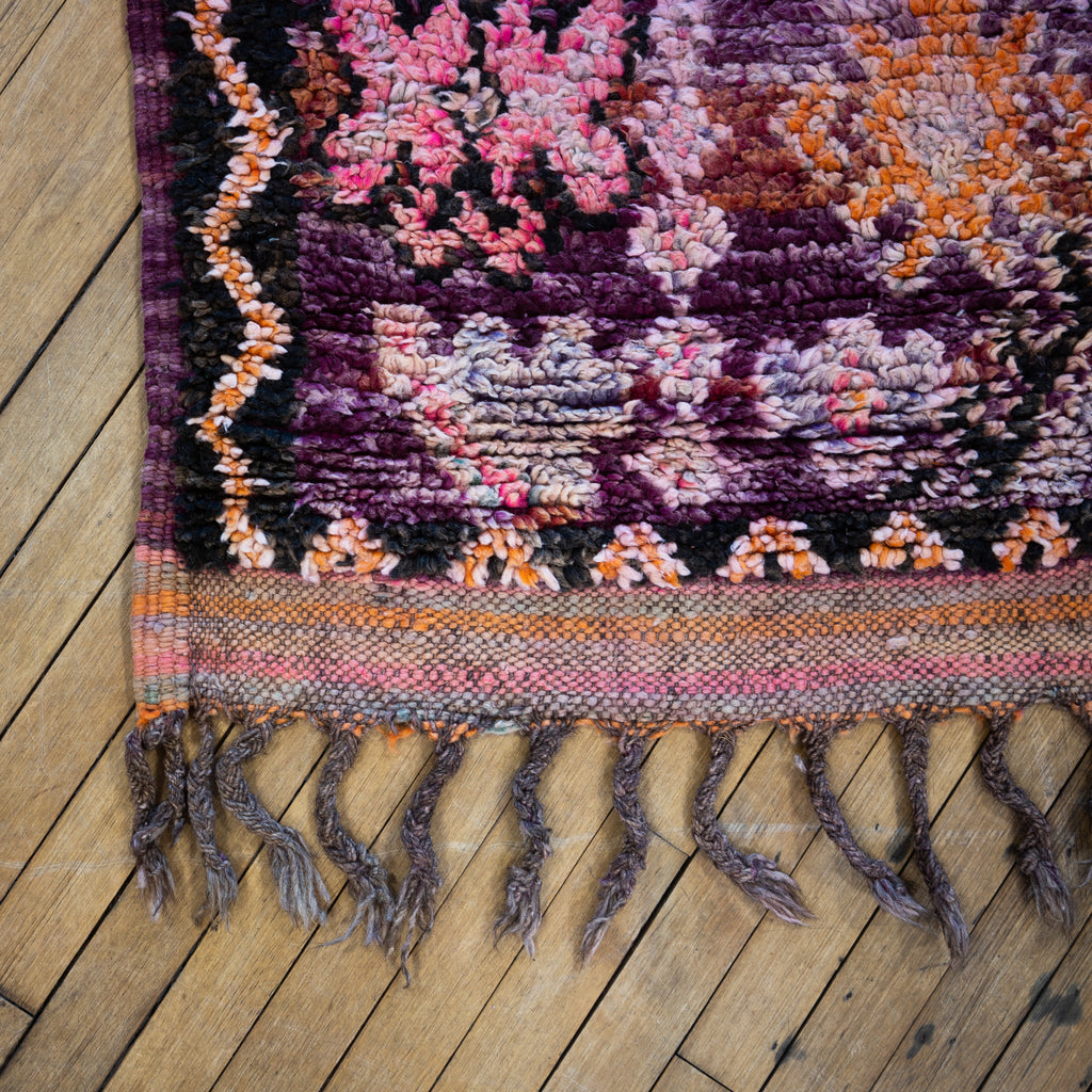 Close up of the fringed corner of a vintage Moroccan Boujad rug with a bold diamond design in shades of purple, orange, and pink. Two diamonds are surrounded by various Berber symbols. A zig zag border surrounds an edge section with repeat Berber symbols.. Wood floor.