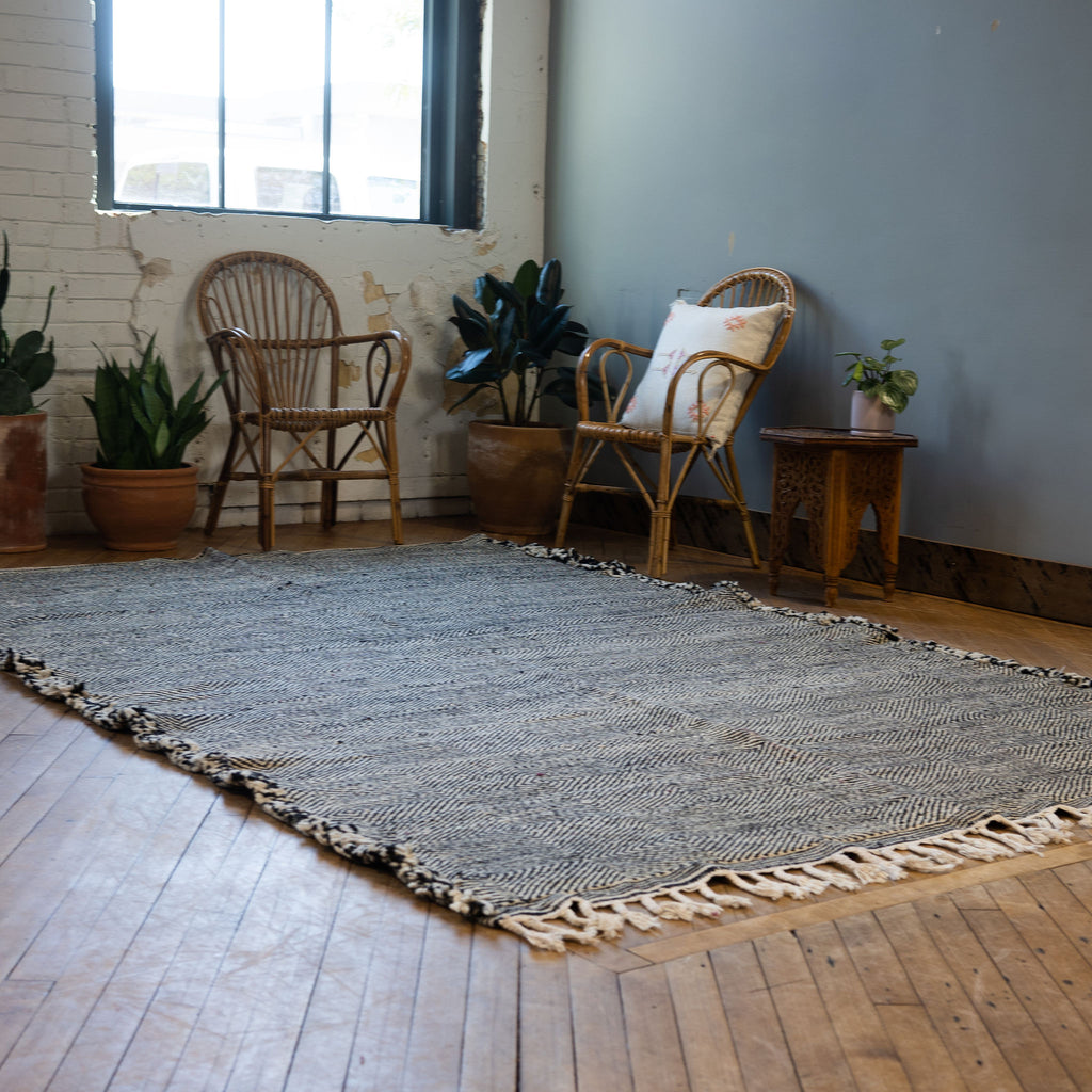 A diagonal view of a flat woven wool Moroccan Zanafi rug with a subtle geometric radiating diamond design in black and white surrounded by two rattan chairs, a side table, and a potted plant. Wood floors.