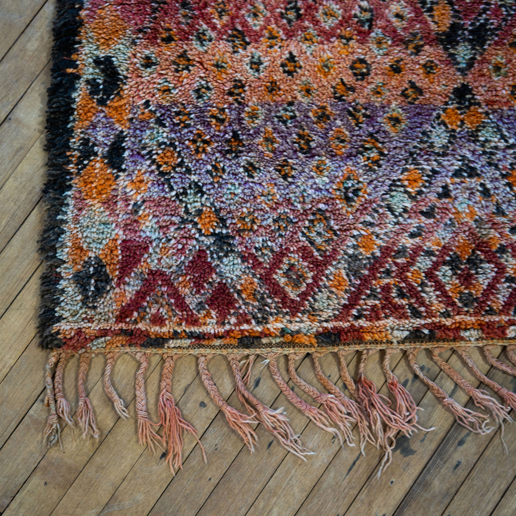 Close up of the fringed corner of a vintage Moroccan Boujad rug with a bold Berber pattern featuring layered diamonds in shades of purple, red, orange, brown, cream, and teal. Wood floor.