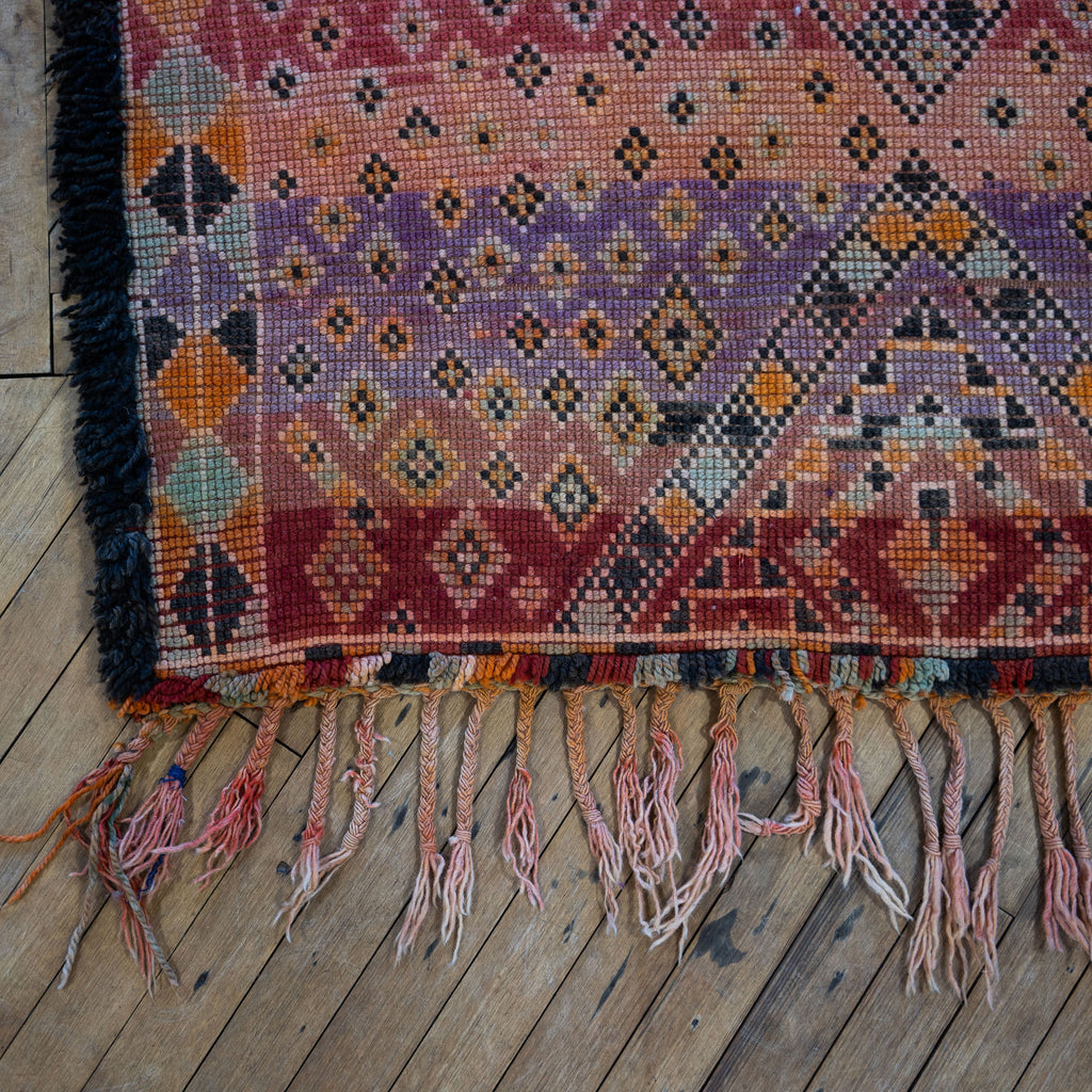 Close up of the summer side or back side fringed corner of a vintage Moroccan Boujad rug with a bold Berber pattern featuring layered diamonds in shades of purple, red, orange, brown, cream, and teal. Wood floor.