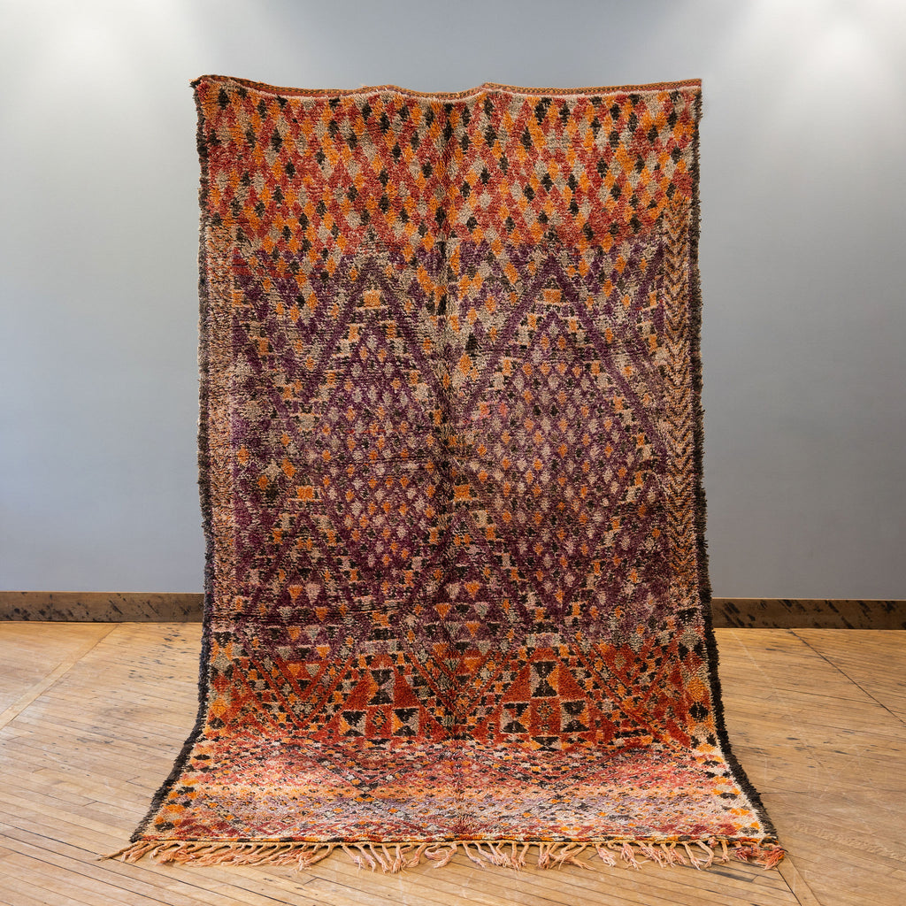 A vintage Moroccan Boujad rug with a bold Berber pattern featuring layered diamonds in shades of purple, red, orange, brown, cream, and teal. Rug is held up against a grey wall and wood floor.