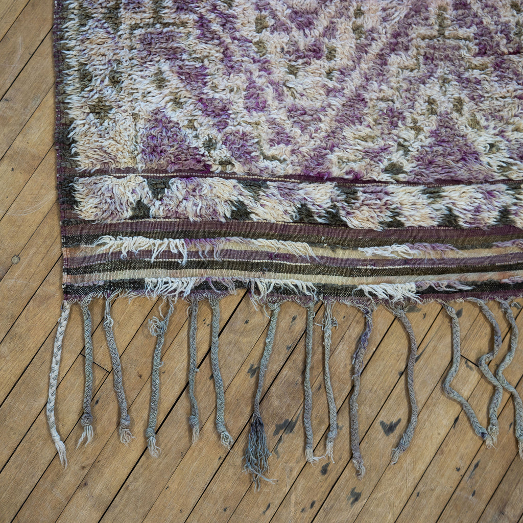 Close up of the fringed corner of a vintage Moroccan Boujad rug with a bold geometric Berber ombre pattern featuring diamonds and zig zags in shades of purple, olive green, cream, and peach. Wood floor.