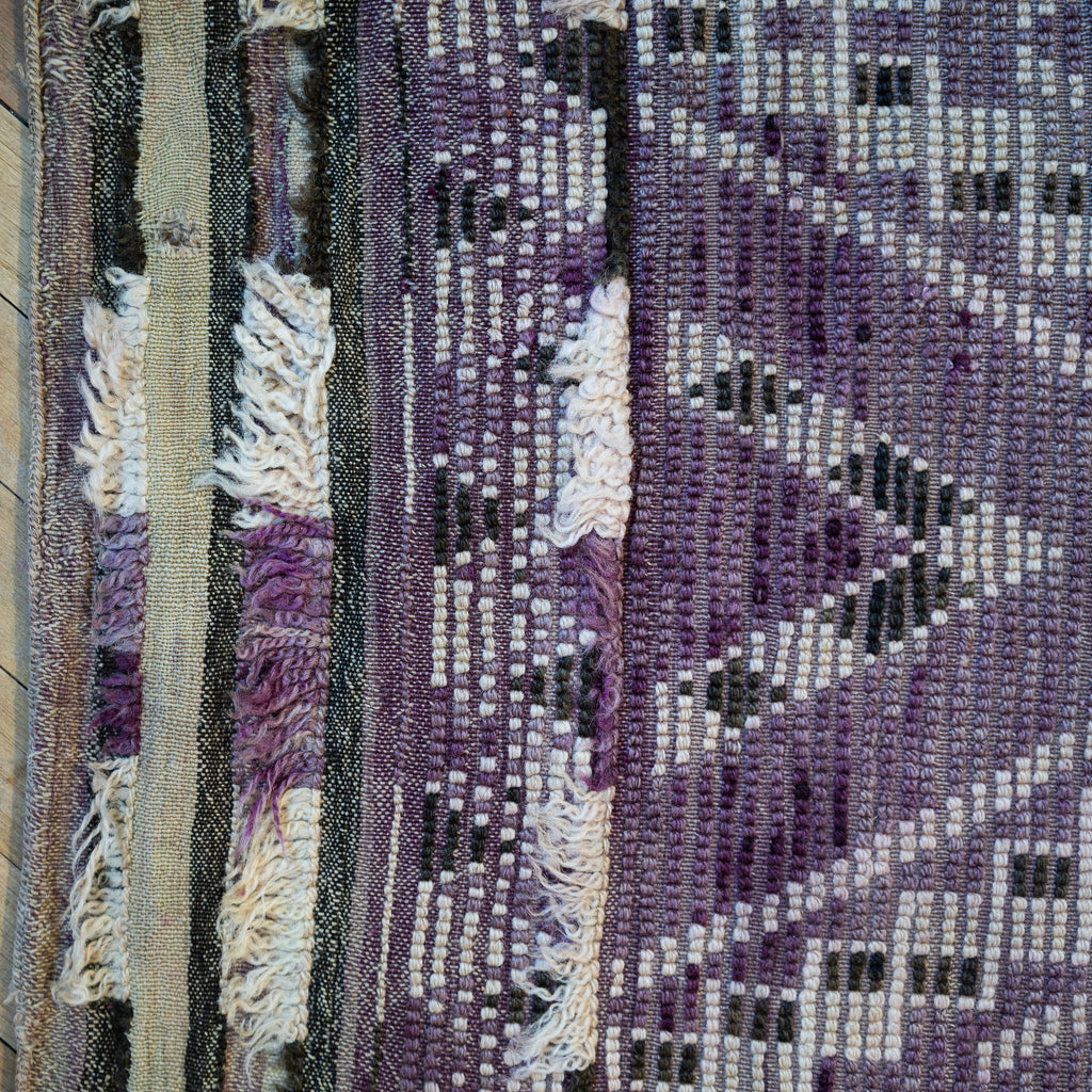 Close up of the summer side or back side of a vintage Moroccan Boujad rug with a bold geometric ombre Berber pattern featuring diamonds and zig zags in shades of purple, olive green, cream, and peach. Wood floor.