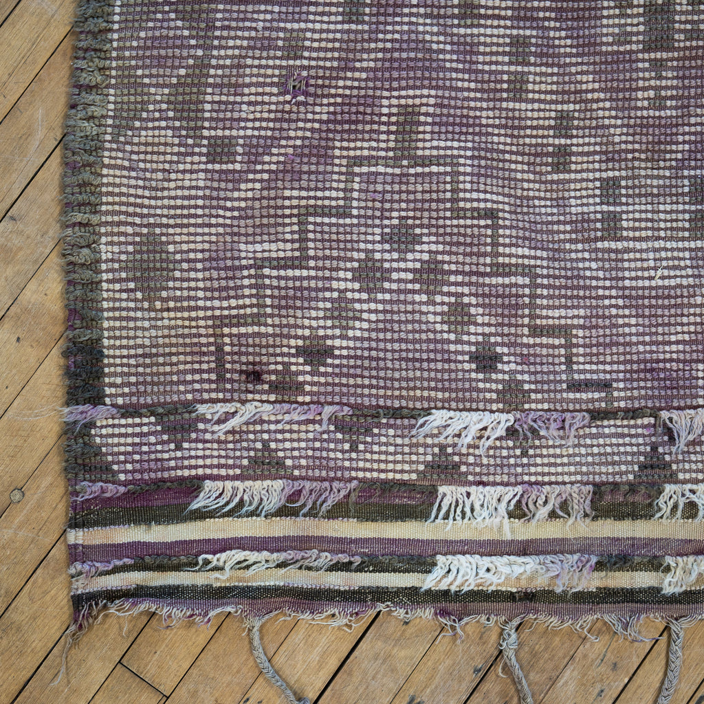 Close up of the summer side or back side fringed corner of a vintage Moroccan Boujad rug with a bold geometric ombre Berber pattern featuring diamonds and zig zags in shades of purple, olive green, cream, and peach. Wood floor.