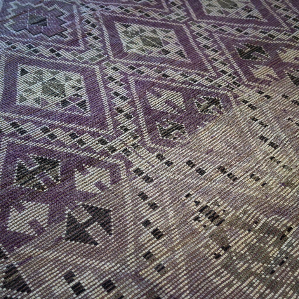 Close up of the summer side or back side of a vintage Moroccan Boujad rug with a bold geometric Berber pattern featuring diamonds and zig zags in shades of purple, olive green, cream, and peach. Wood floor.