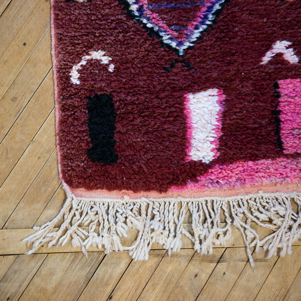 Close up of the fringed corner of a vintage Moroccan Boujad runner with a bold modern abstract design in maroon, pink, black, purple, tan, and cream. Various shapes and lines balance each other out in the abstract layout. Wood floor.