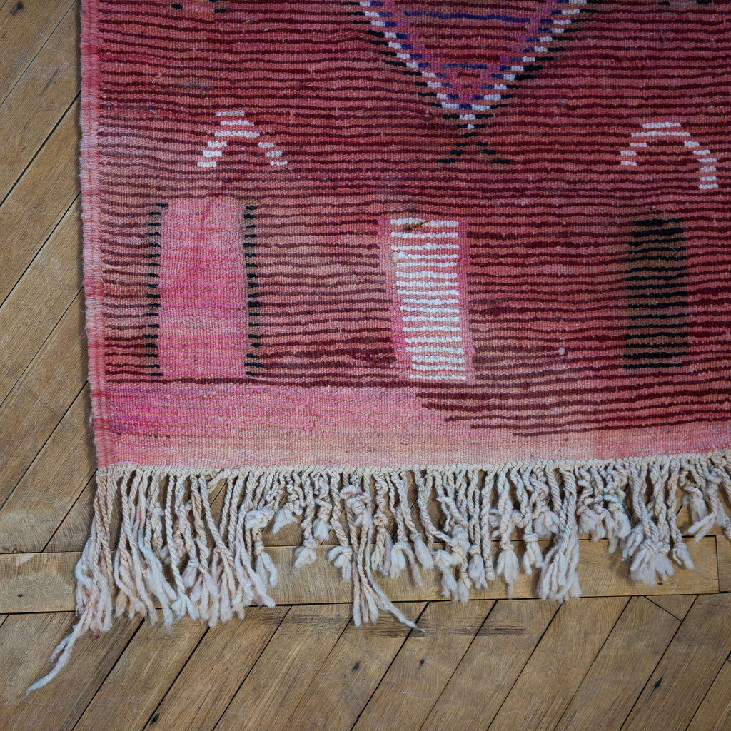 Close up of the summer side or back side fringed corner of a vintage Moroccan Boujad runner with a bold modern abstract design in maroon, pink, black, purple, tan, and cream. Various shapes and lines balance each other out in the abstract layout. Wood floor.