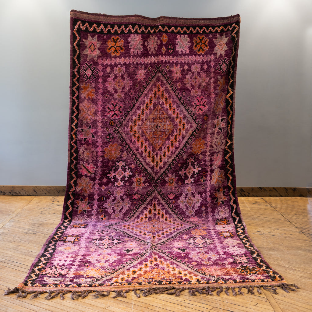 A vintage Moroccan Boujad rug with a bold diamond design in shades of purple, orange, and pink. Two diamonds are surrounded by various Berber symbols. A zig zag border surrounds an edge section with repeat Berber symbols. Rug is held up against a grey wall and wood floor.