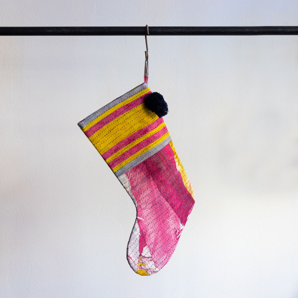 A holiday stocking that is handmade and embroidered with the traditional Kantha stitch. Recycled sari fabric in bright pink, yellow, white and gray join black poms at the hanging loop. Hanging from a brass s-hook from a black bar in front a white background.