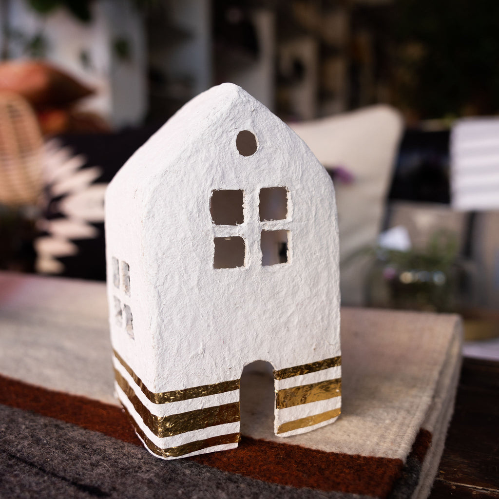 White and gold leaf cotton mache house. Gold leaf stripes run along the bottom of the house. Sits on a woven rug in front of a blurry background.