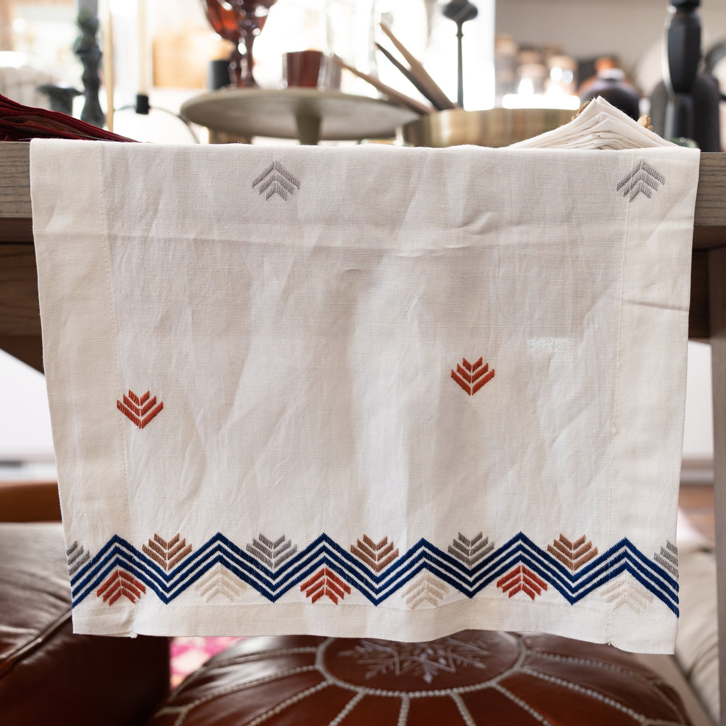 Cream linen table runner with geometric arrow design throughout and zig-zag + arrow combo on edges. Blue, grey, cream, tan, and orange embroidery.