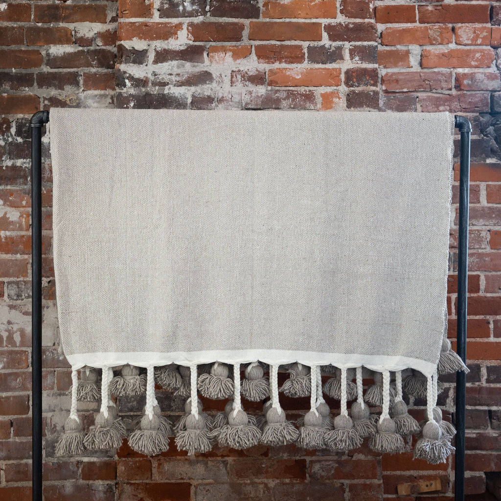 Woven cotton taupe colored blanket with poms on the edge hangs over a black rod in front of a brick wall.