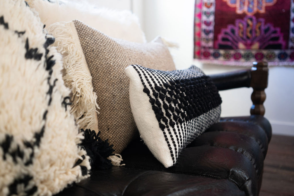 Group of textural black and cream and tan wool pillows on a black leather sofa in front of a colorful rug.