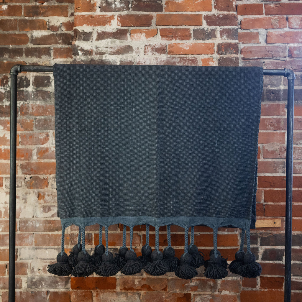 Woven cotton navy colored blanket with poms on the edge hangs over a black rod in front of a brick wall.