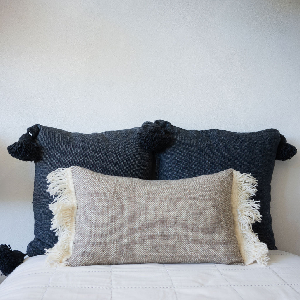 Tan and cream wool tweed pillow sits in front of two square woven cotton navy blue pillows with big poms at each corner.