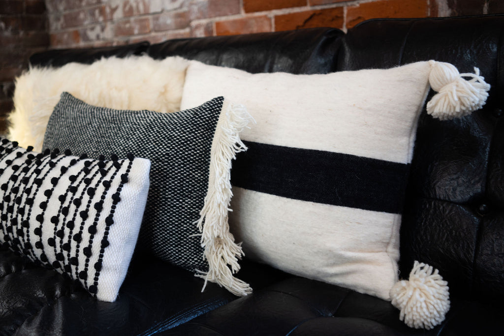 Group of textured black and cream wool pillows on a black leather couch in front of a brick wall. 
