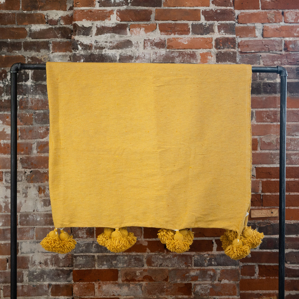 Woven mustard colored blanket with poms on the edge hangs over a black rod in front of a brick wall.