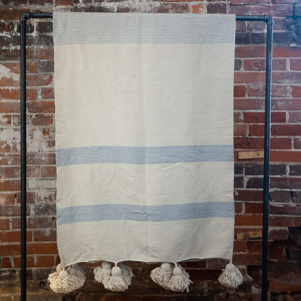 Woven cotton blanket with poms on edge. Natural cream is broken up by varying widths of gray stripes. Blanket hangs over a black rod in front of a brick wall. 