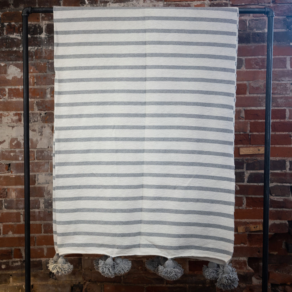Woven cotton gray and cream striped blanket with poms on the edge hangs over a black rod in front of a brick wall.
